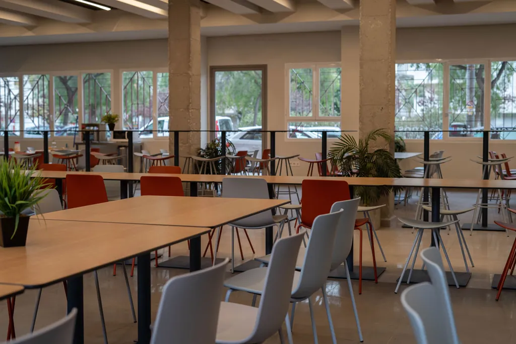 Comedor de la residencia con grandes ventanales, mesas de madera y sillas de colores