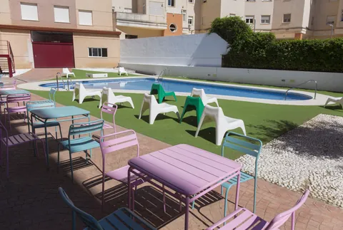 Patio de la residencia con la piscina, césped y mesas de colores al sol